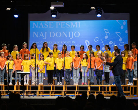 Otroški pevski zbor nastopa na prireditvi 'Naše pesmi naj donijo' v oranžnih in rumenih majicah na odru.