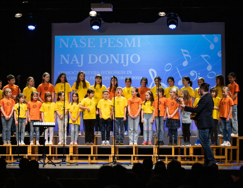 Otroški pevski zbor nastopa na prireditvi 'Naše pesmi naj donijo' v oranžnih in rumenih majicah na odru.